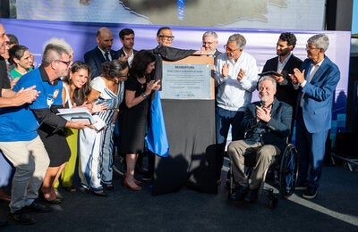 Grupo de autoridades, pesquisadores e participantes reunidos ao redor da placa de inauguração durante a cerimônia de reabertura do Centro Cultural do Ministério da Saúde, no Rio de Janeiro. Algumas pessoas aplaudem enquanto a placa comemorativa é descerrada; em primeiro plano, um homem em cadeira de rodas sorri e participa do momento.