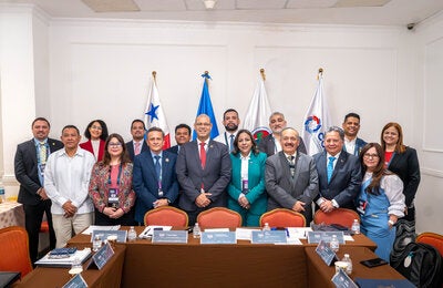 Asistentes al foro posan en el salón del evento. Banderas de las instituciones figuran al fondo.