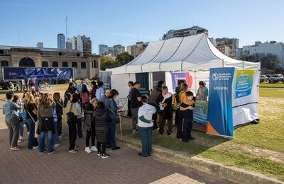 Un grupo de personas espera frente a un gazebo blanco, con banderas y banners de OPS, para aplicarse sus vacunas en la Feria del Libro.