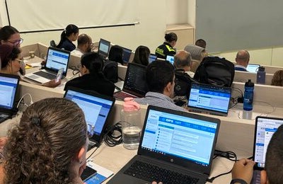 personas en salón de clases tipo auditorio, usando computador. Plataforma SIMEX visible en la pantalla.