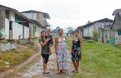Tres mujeres y un niño posan en una calle de una comunidad amazónica rural, rodeados de viviendas de madera y áreas verdes en un día lluvioso.