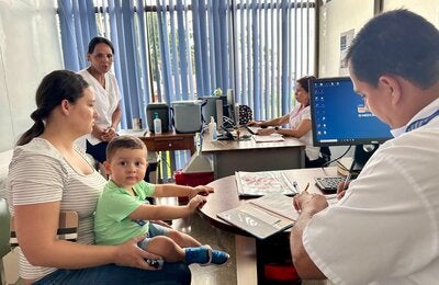 Persona adulta con niño pequeño en brazos durante una consulta en un centro de salud, mientras personal sanitario atiende y registra información en una computadora.