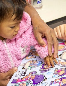Foto de niña con su mamá a la espera de ser vacuna mientras juega con un rompecabezas sobre vacunas