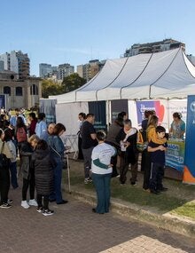 Un grupo de personas espera frente a un gazebo blanco, con banderas y banners de OPS, para aplicarse sus vacunas en la Feria del Libro.