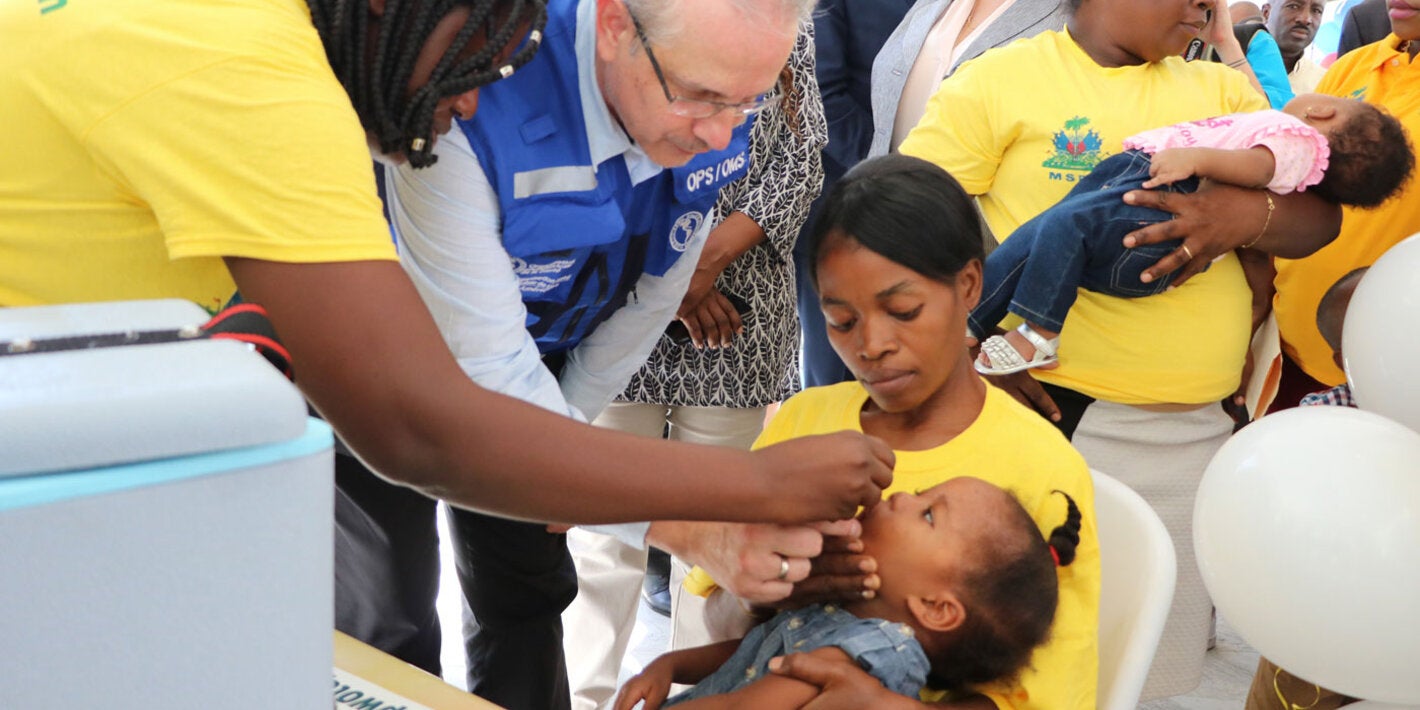Le Représentant de l'OPS/OMS en Haïti, le Dr Luis Felipe Codina administre un vaccin Polio Oral à un enfant sous le regard de la Ministre de la Sante Publique