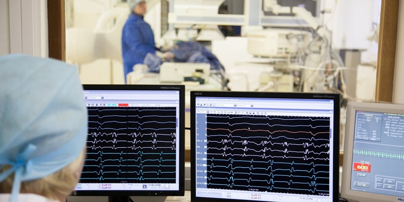 Photo of the back of the head of a healthworker, behing him three screens showing paralel lines that indicates the heart function. Behind the screens there is a big window leading to a room with health equipment where another health worker is accompanying a patient laid on the machine