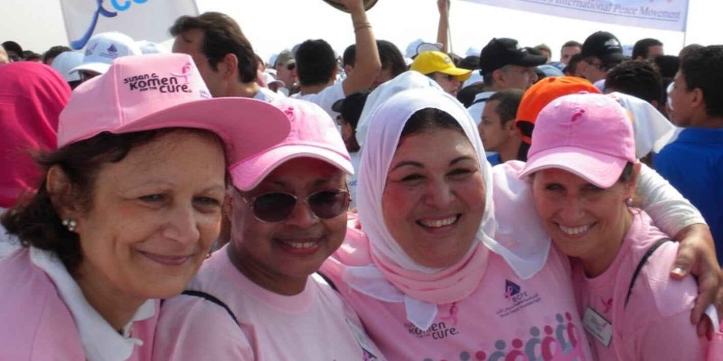 A group of four women dressed in pink t shirts embrace each other smiling. In the background other people is gathering with some banners on a breast cancer awarenes event