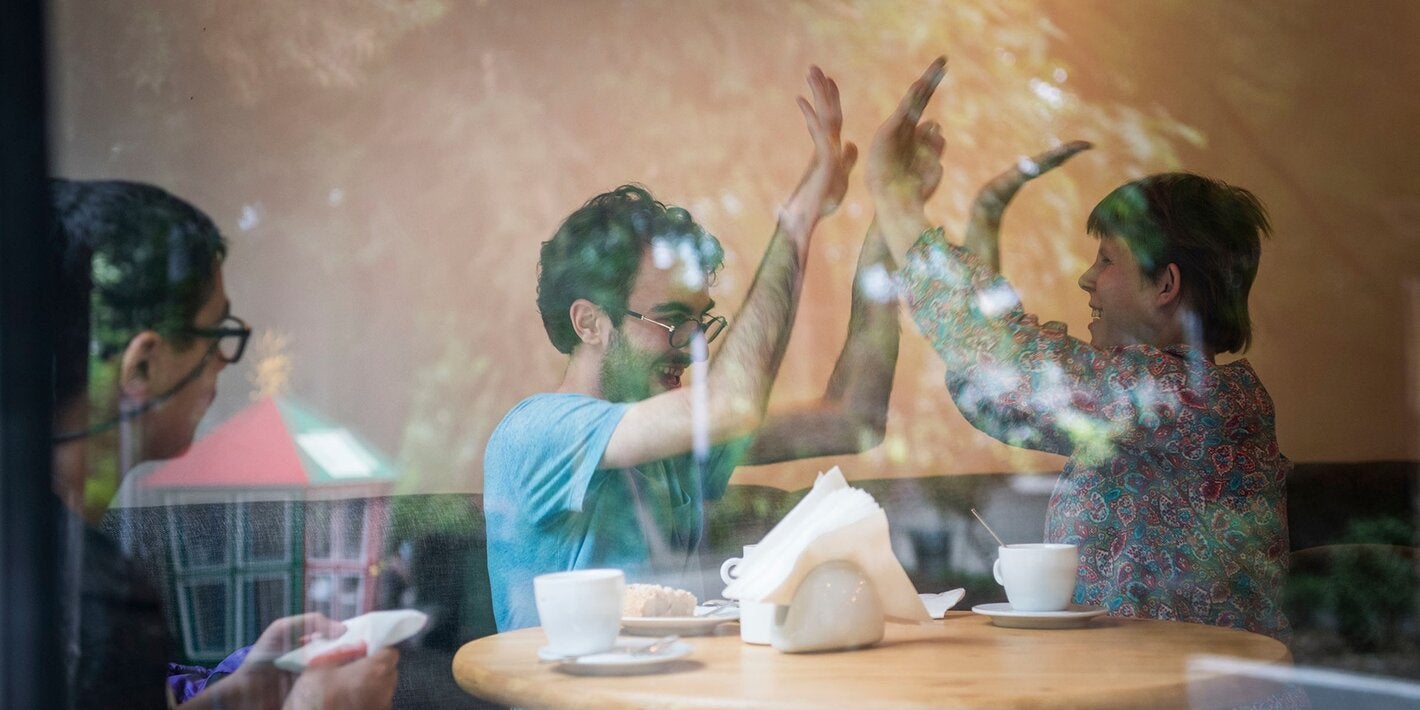 A man and a woman sitting next to a coffee table shake both hands with their arms up, while a third man watches the scene from the left