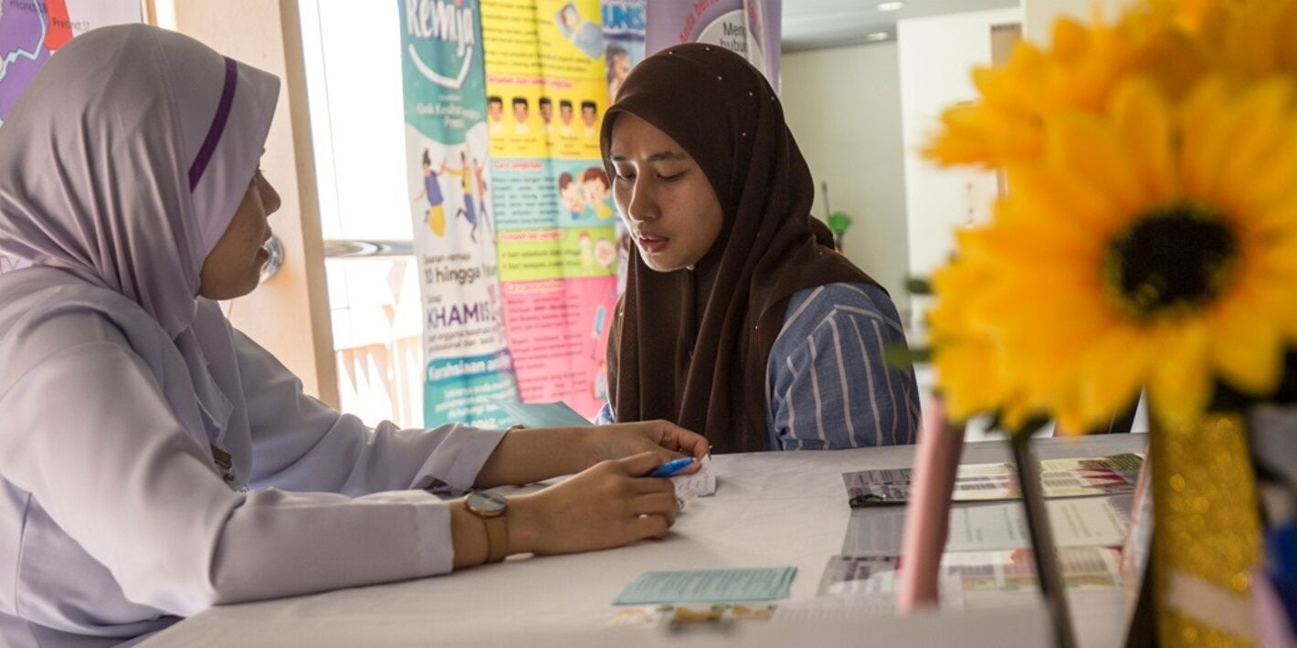 Photo of two women in health care facility; one is the patient, the other the health provider. Both have their heads covered with a scarf 