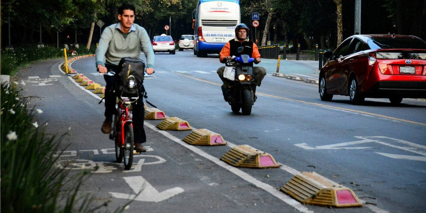 Ciclista circulando por una ciclovía protegida, en la calle circula de frente una motocicleta y dos vehículos a motor en sentido contrario