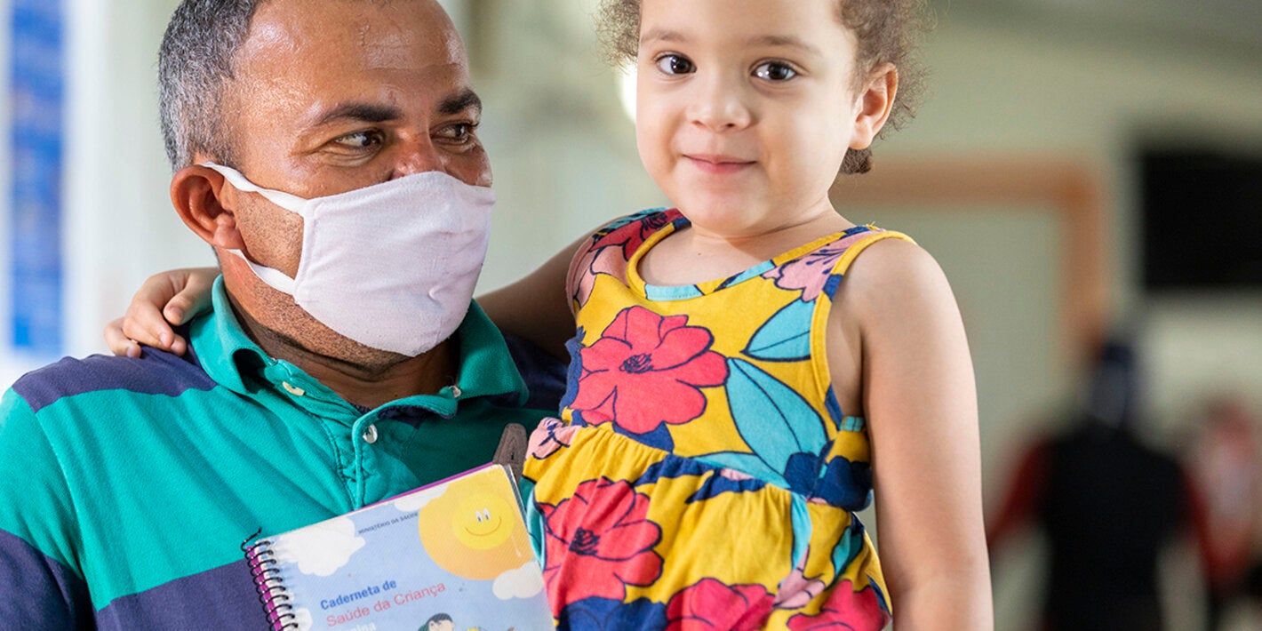 Criança sorrindo e olhando para a câmera, no colo do pai, após ter se vacinado contra pólio