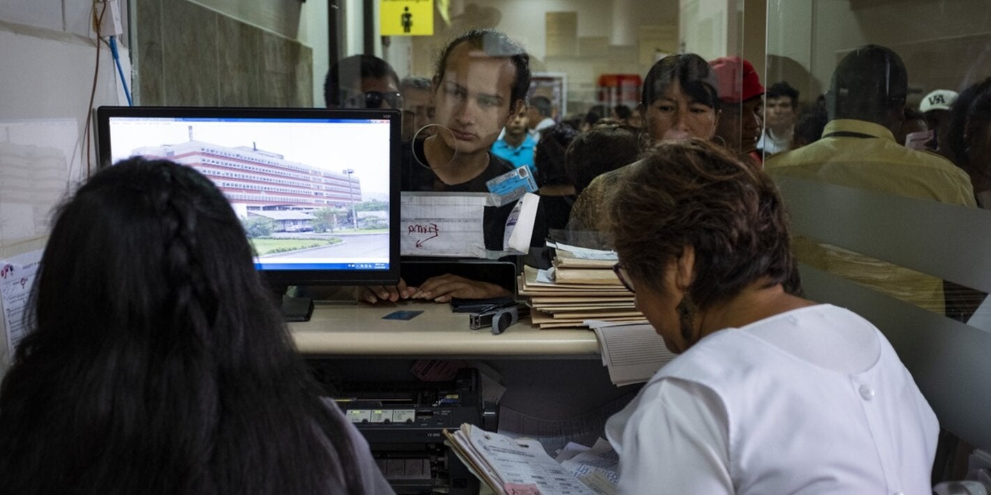Image of the registration area in a hospital, where two health workers are attending a line of people in front of her