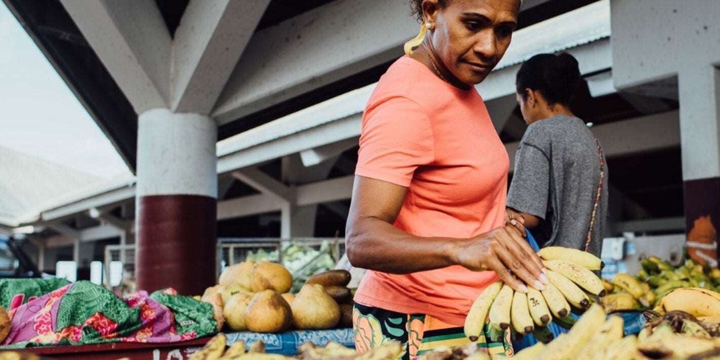 Photo of a middle aged woman dressed with an orange t-shirt grabbing a bunch of bananas on a market fruit stall