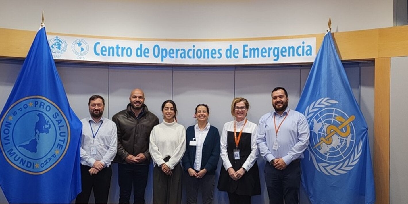 Personas (hombres y mujeres) posan a la cámara en el Centro de Operaciones de Emergencias de la OPS. A ambos lados, están las banderas de la OPS y la OMS.