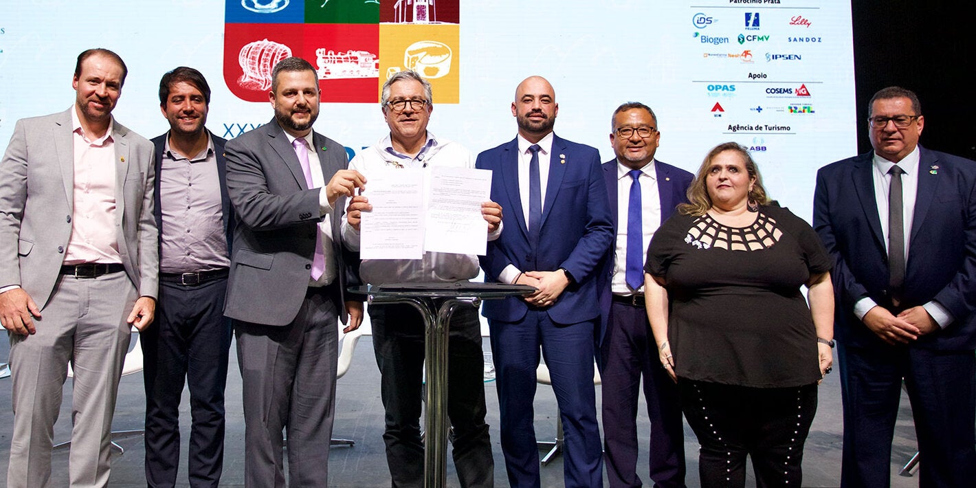 O Ministro da Saúde do Brasil, Alexandre Padilha, segura documentos assinados por ele. Na foto, ao lado dele estão o presidente do Conasems e outras autoridades.