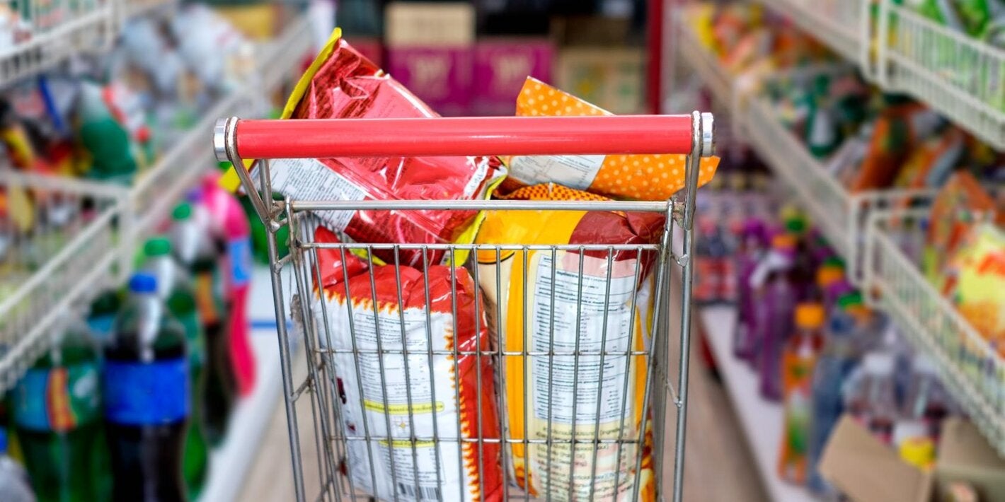Photo of a supermarket cart filled with huge bags of chips, in the middle of two supermarket aisles with more ultraprocessed products