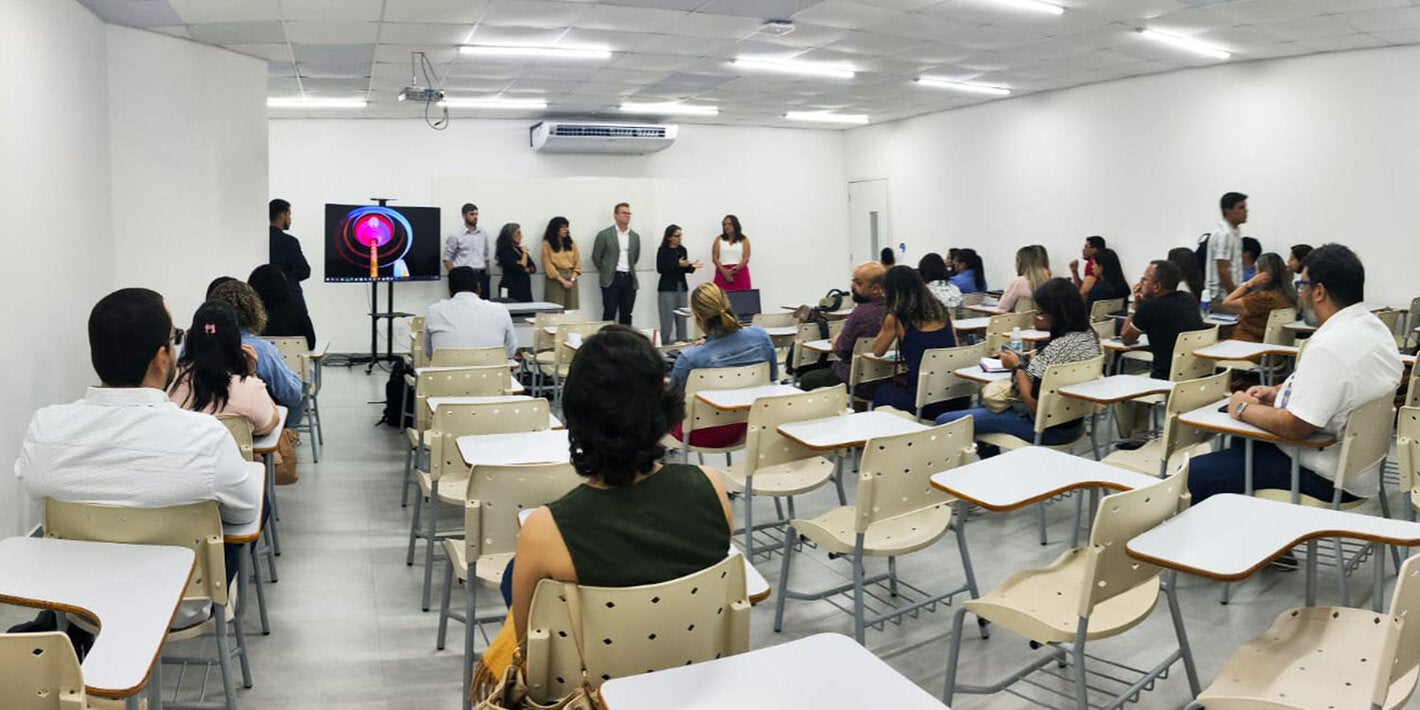 Profissionais sentados em mesas de sala de aula, ouvindo palestra da equipe da OPAS e parceiros