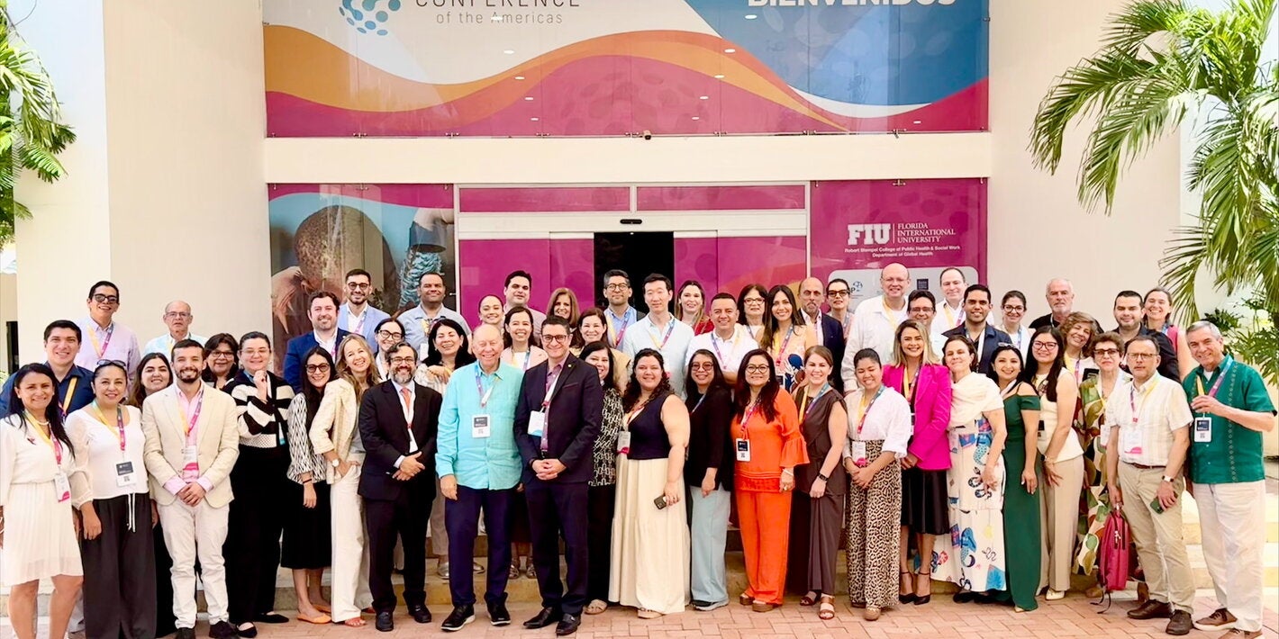 Los participantes del taller “Evidencia y herramientas disponibles para la eliminación del cáncer cervicouterino en las Américas” posan para una foto grupal frente a la entrada de un edificio. En el fondo, se puede ver un cartel que dice "GLOBAL HEALTH CONFERENCE of the Americas" y "BIENVENIDOS". También hay logotipos de FIU (Florida International University) y otros elementos decorativos. Los asistentes están vestidos con ropa formal y llevan gafetes colgados al cuello.