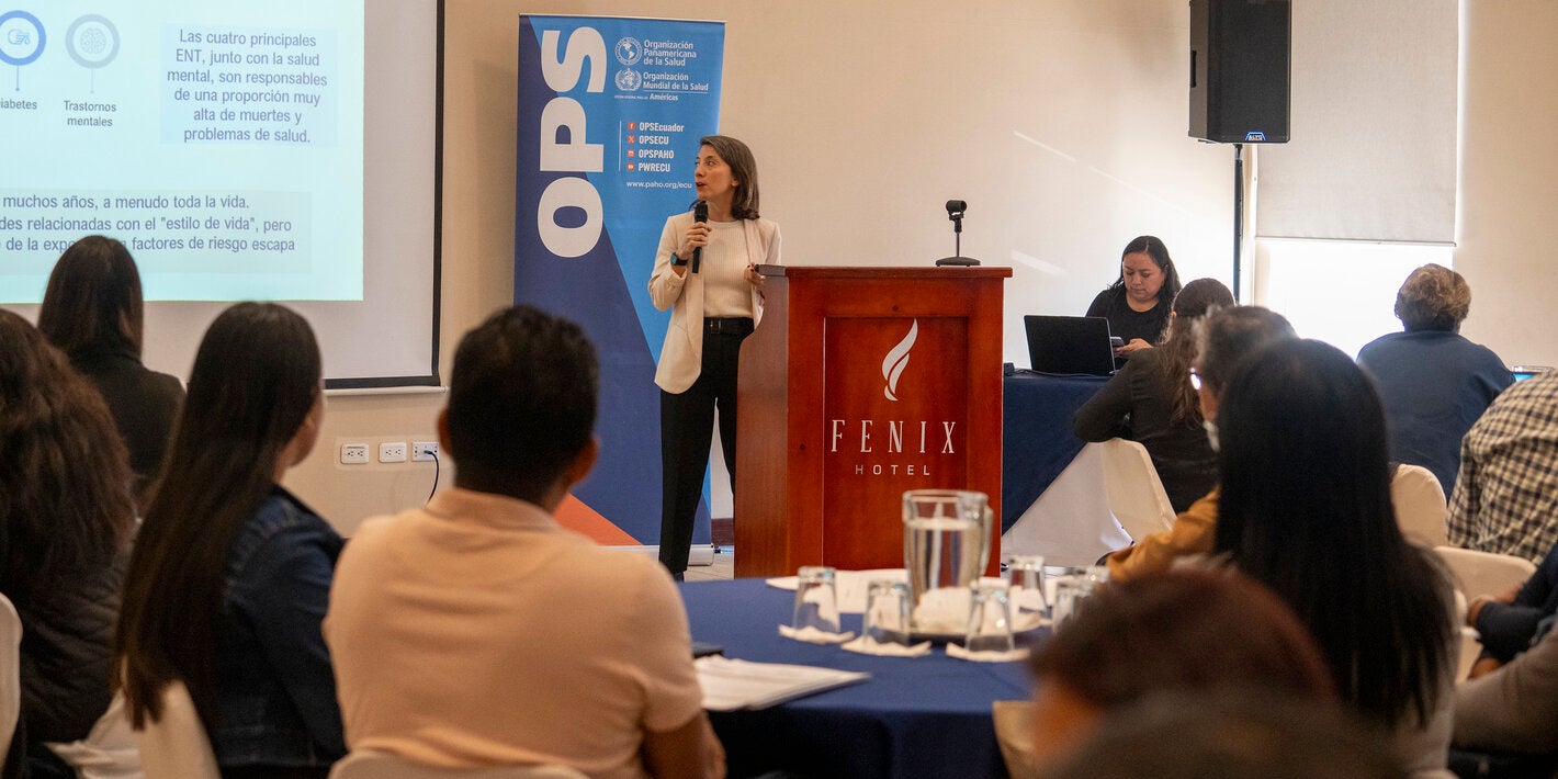 Sesión de entrenamiento de la encuesta STEPS en Quito, Ecuador (8–10 de septiembre de 2025), con una persona presentando frente a un atril del Fenix Hotel, acompañada de un banner de la OPS. Se proyecta una diapositiva sobre enfermedades no transmisibles, mientras la audiencia escucha desde mesas redondas con manteles azules.
