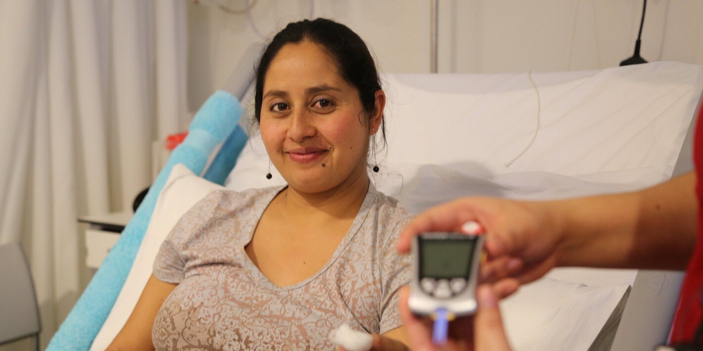 Pregnant woman sitting on a hospital bed holding a piece of cotton after a blood sample, while another hand in the foreground displays a blood glucose meter