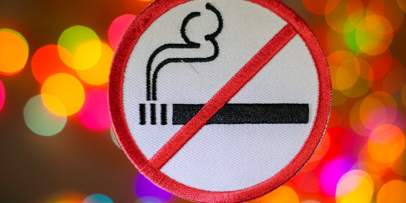 Round sign with a red border showing a black cigarette with smoke inside a red diagonal line, indicating no smoking. The background is blurred with colorful bokeh lights