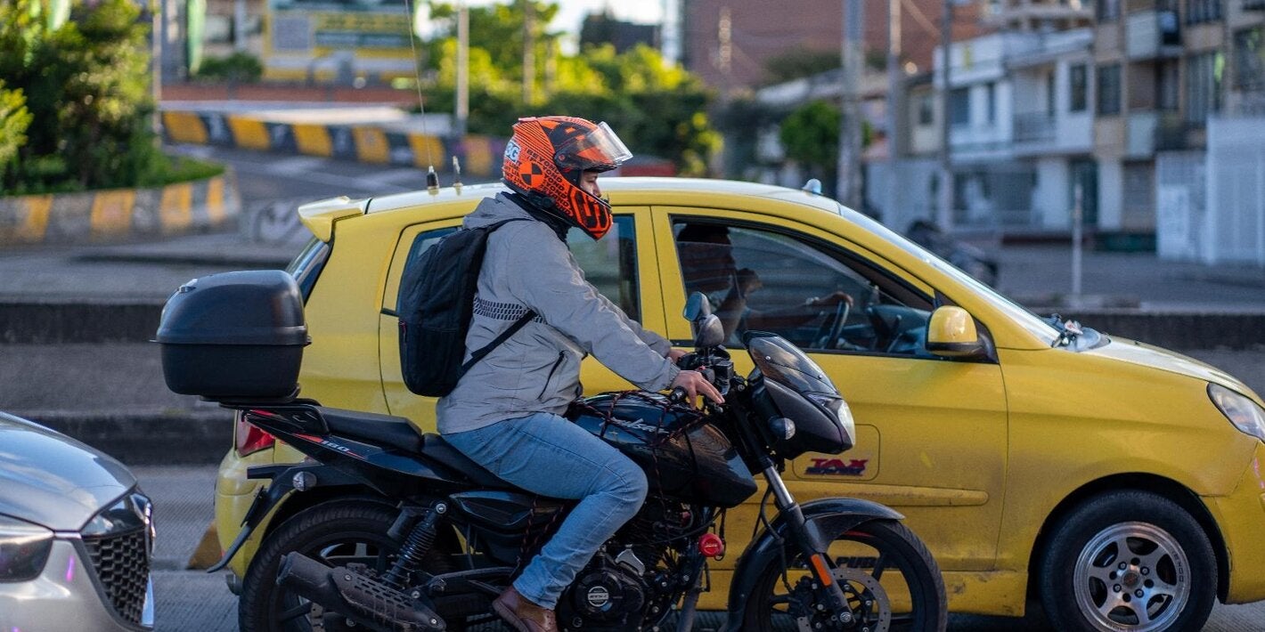 Motociclista con casco rojo, circulando en una avenida urbana, con un vehículo amarillo a su lado izquierdo