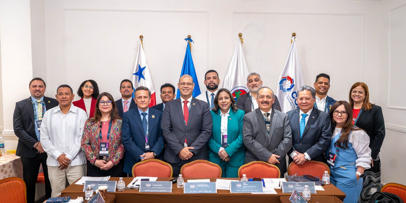 Asistentes al foro posan en el salón del evento. Banderas de las instituciones figuran al fondo.