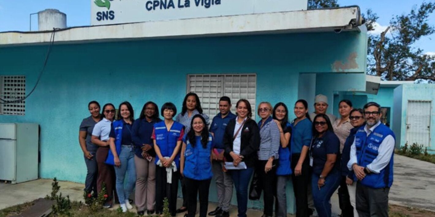 Grupo de trabajadores de salud del centro de atención primaria La Vigía y expertos de OPS posan frente al centro de salud, que tiene las paredes pintadas de verde aguamarina, con un cielo azul con nubes dispersas y un frente de hierba