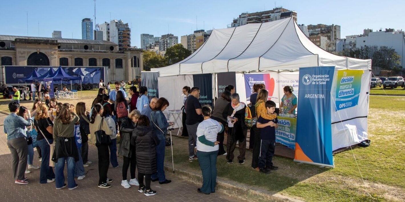 Un grupo de personas espera frente a un gazebo blanco, con banderas y banners de OPS, para aplicarse sus vacunas en la Feria del Libro.