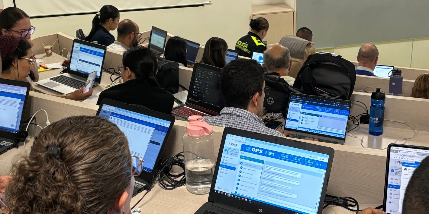 personas en salón de clases tipo auditorio, usando computador. Plataforma SIMEX visible en la pantalla.