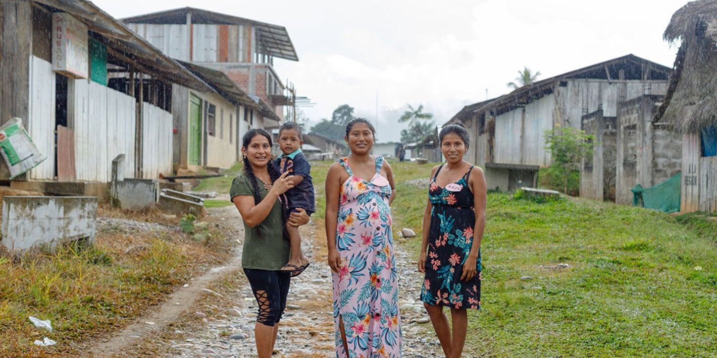 Tres mujeres y un niño posan en una calle de una comunidad amazónica rural, rodeados de viviendas de madera y áreas verdes en un día lluvioso.