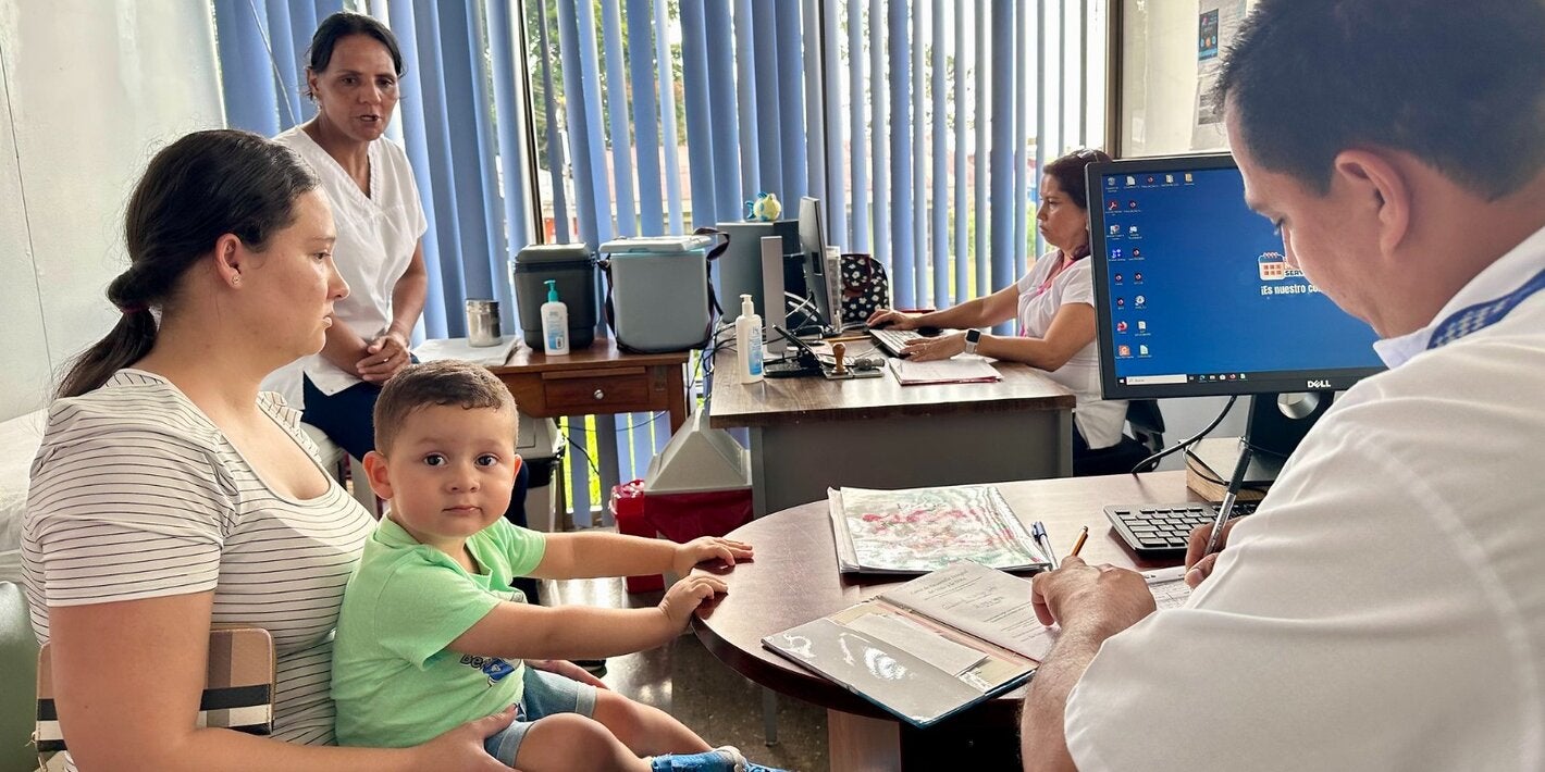 Persona adulta con niño pequeño en brazos durante una consulta en un centro de salud, mientras personal sanitario atiende y registra información en una computadora.
