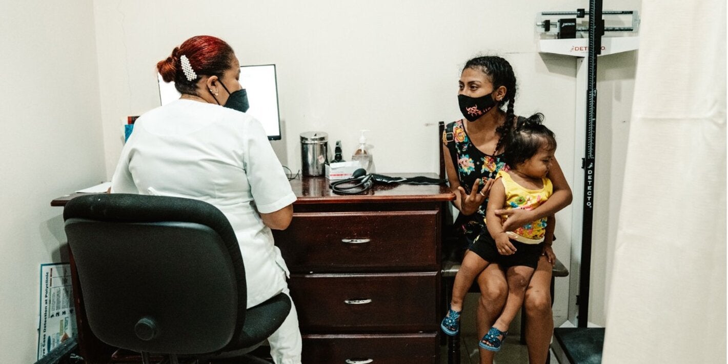 Health personnel seated in a consultation room speaks with an adult holding a young child in their arms.