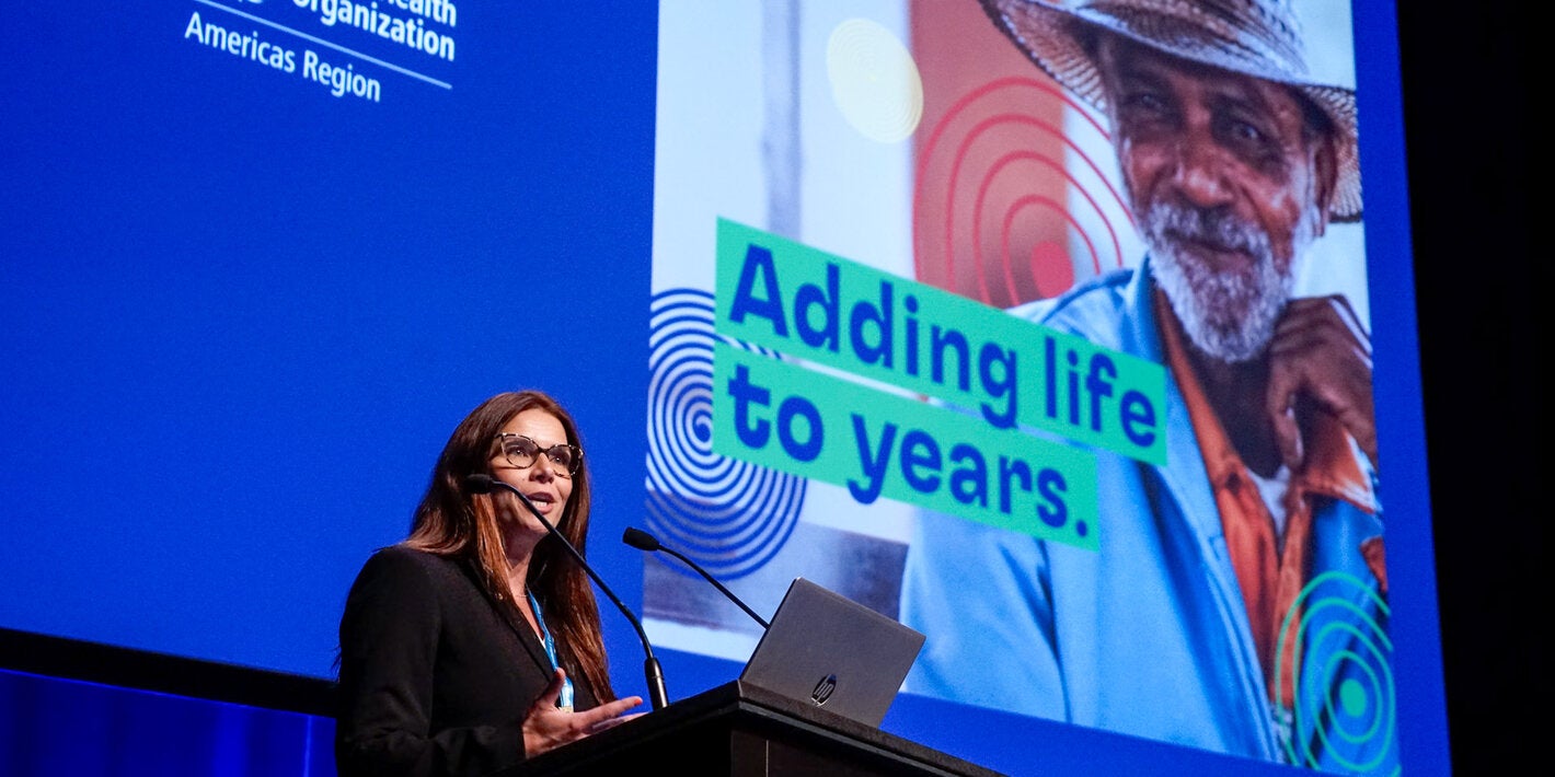 Woman on podium giving speech with presentation on a big screen behind her, with the Decade of Healthy Aging in the Americas card with slogan that reads "Adding life to years"