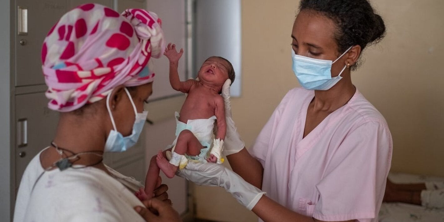 doctor with mask holding newborn baby in clinic in front of its mother