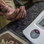 Close up of a glucometer and in the back a hand holding the hand of a patient