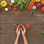 Photo of a brown table seen from above with a couple of hands holding spices recipients and framed by an array of vegetables of different colors