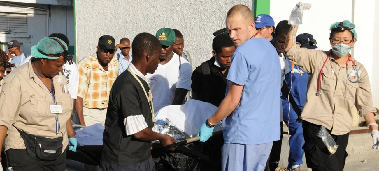 Health personnel and others carrying a patient on a stretcher... one responder is seen holding the IV bag.