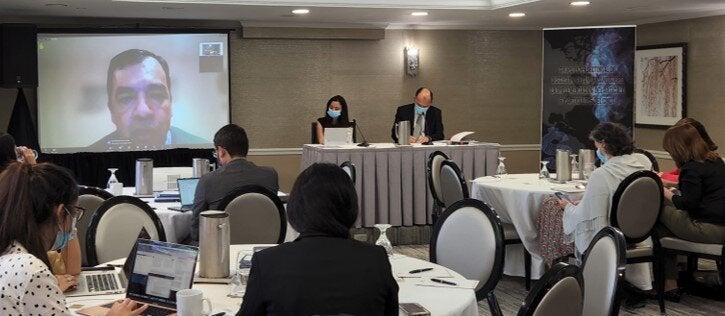 Hybrid meeting with people distributed on tables with safe distance while a man is talking from a screen