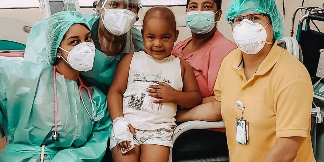 Foto de un grupo de trabajadores sanitarios con máscaras y gorros protectores, rodeando a un paciente con cáncer, un niño calvo de unos 6 años, en un entorno hospitalario