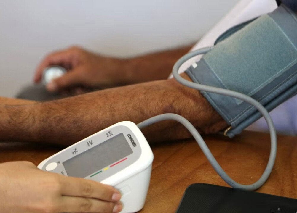A person is having their blood pressure measured using an automatic blood pressure monitor. The person's arm is resting on a table with a cuff wrapped around the upper arm, connected to the monitor by a tube. Another hand is holding the monitor, which displays readings on its screen.
