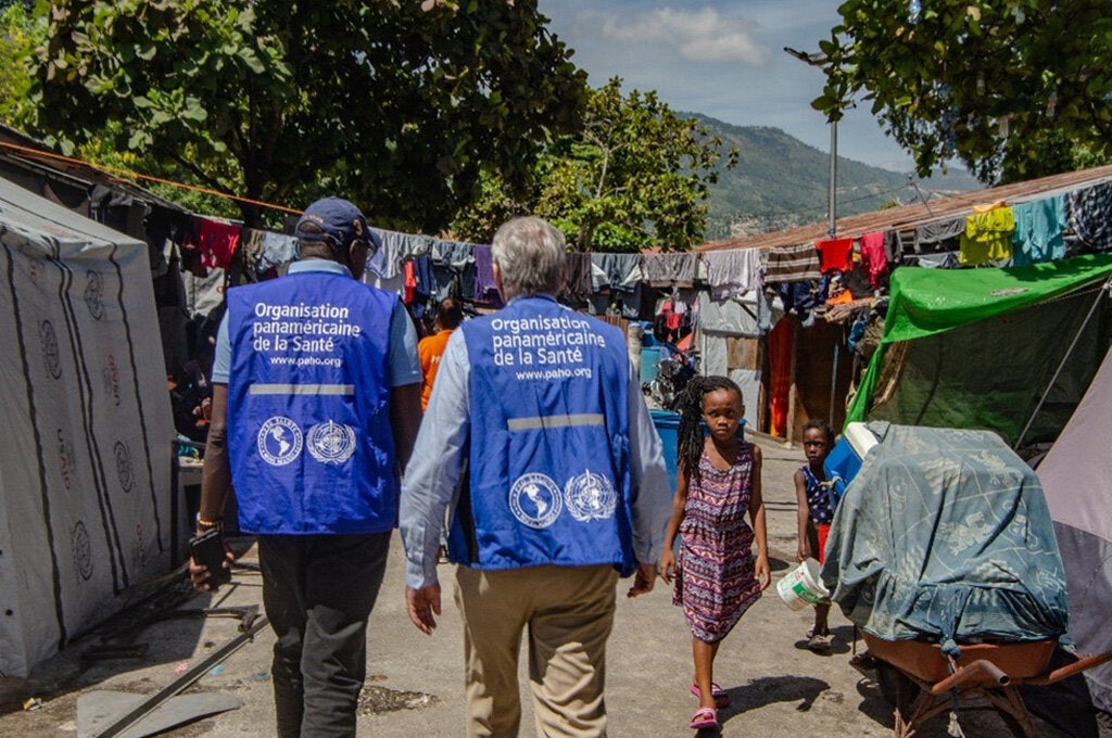 Two PAHO support staff in vests, walking away from camera, in outdoor camp.
