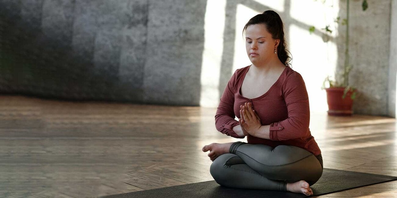 A young woman with Down syndrome is practicing yoga on a yoga mat. She wears a red top and grey yoga pants. Her legs are crossed and her hands are together by the sternum in a "prayer" pose. She gazes down to the ground a few feet in front of her as if in meditation. The room behind her is empty with a wooden floor and a concrete wall and a single potted plant that is climbing up the wall.