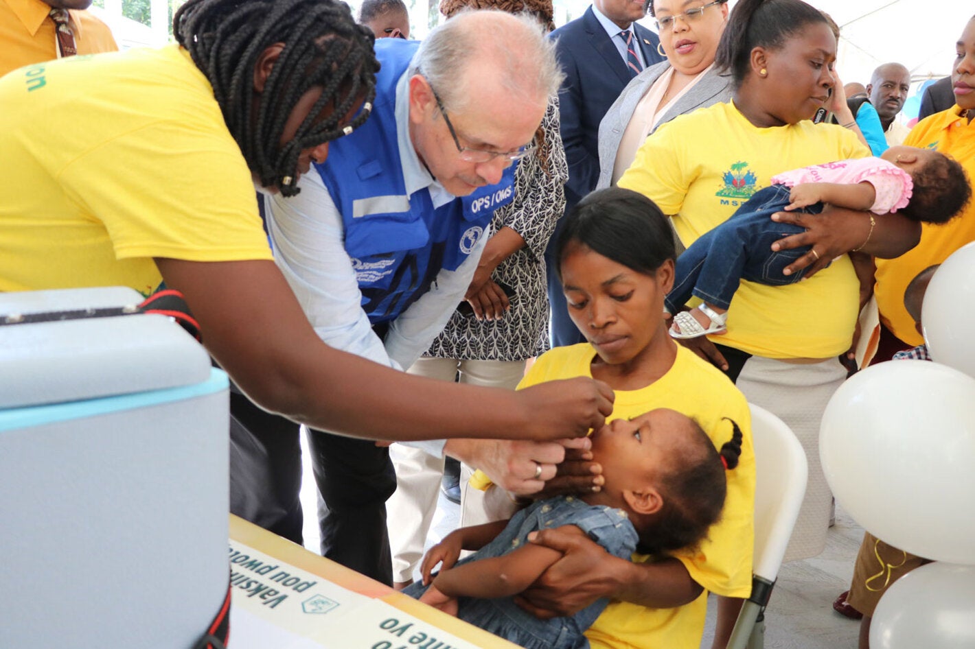 Le Représentant de l'OPS/OMS en Haïti, le Dr Luis Felipe Codina administre un vaccin Polio Oral à un enfant sous le regard de la Ministre de la Sante Publique