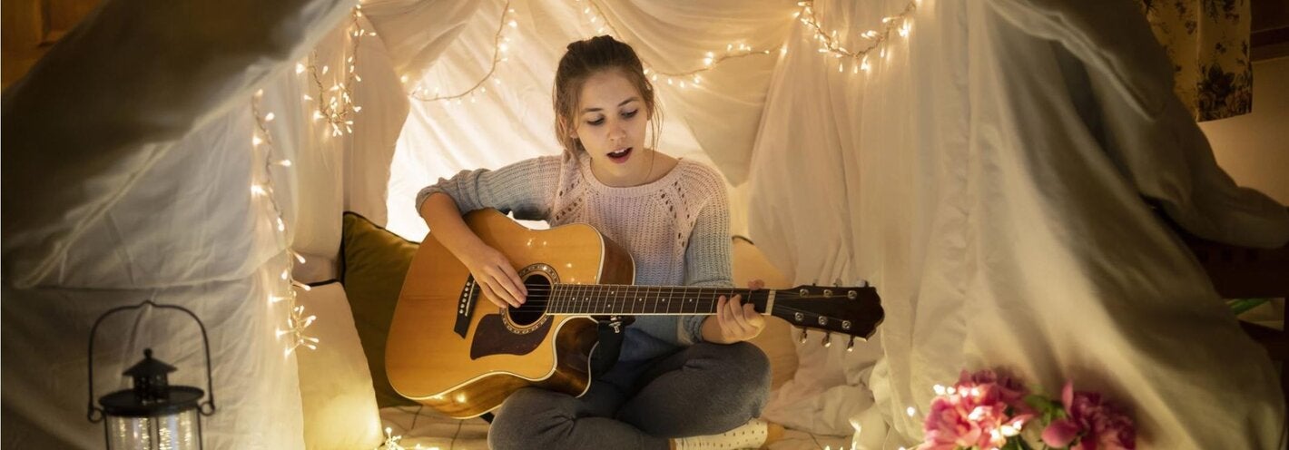Girl playing a guitar under a tent made of cloth illuminated by a string of ligths