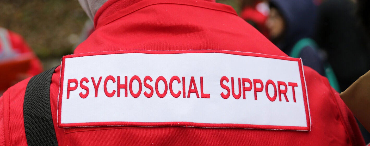 Photo of the back of a healthcare worker dressed with a red jacket with the sign "Psychosocial support" in red typography over white background. Only part of the back is seen, from the top of the shoulders to the mid part of the back