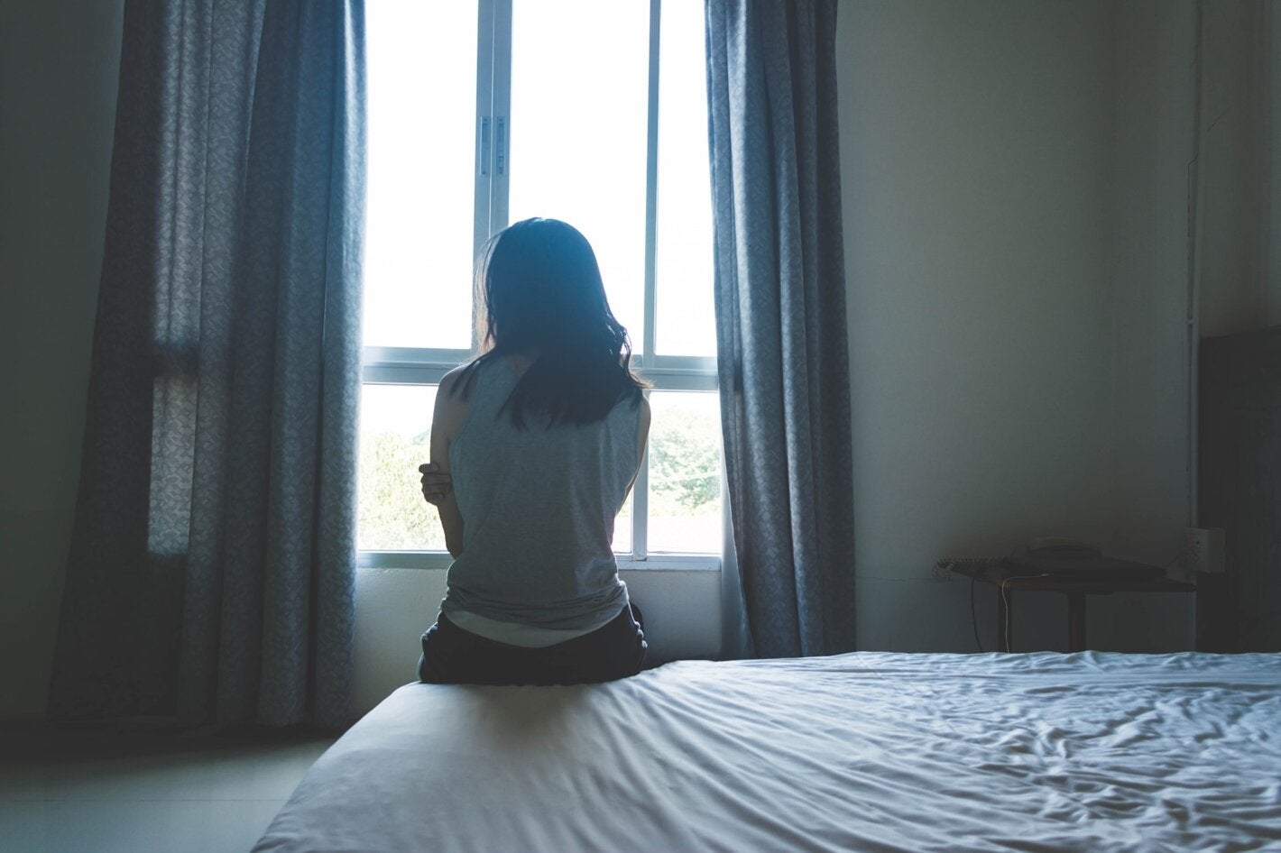 Mujer en la penumbra de una habitación, sentada sobre una cama de espaldas a la cámara, mirando hacia una ventana