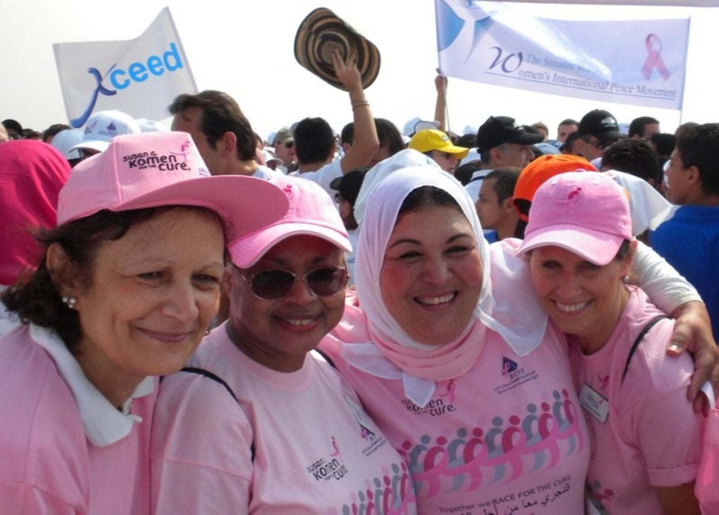 A group of four women dressed in pink t shirts embrace each other smiling. In the background other people is gathering with some banners on a breast cancer awarenes event