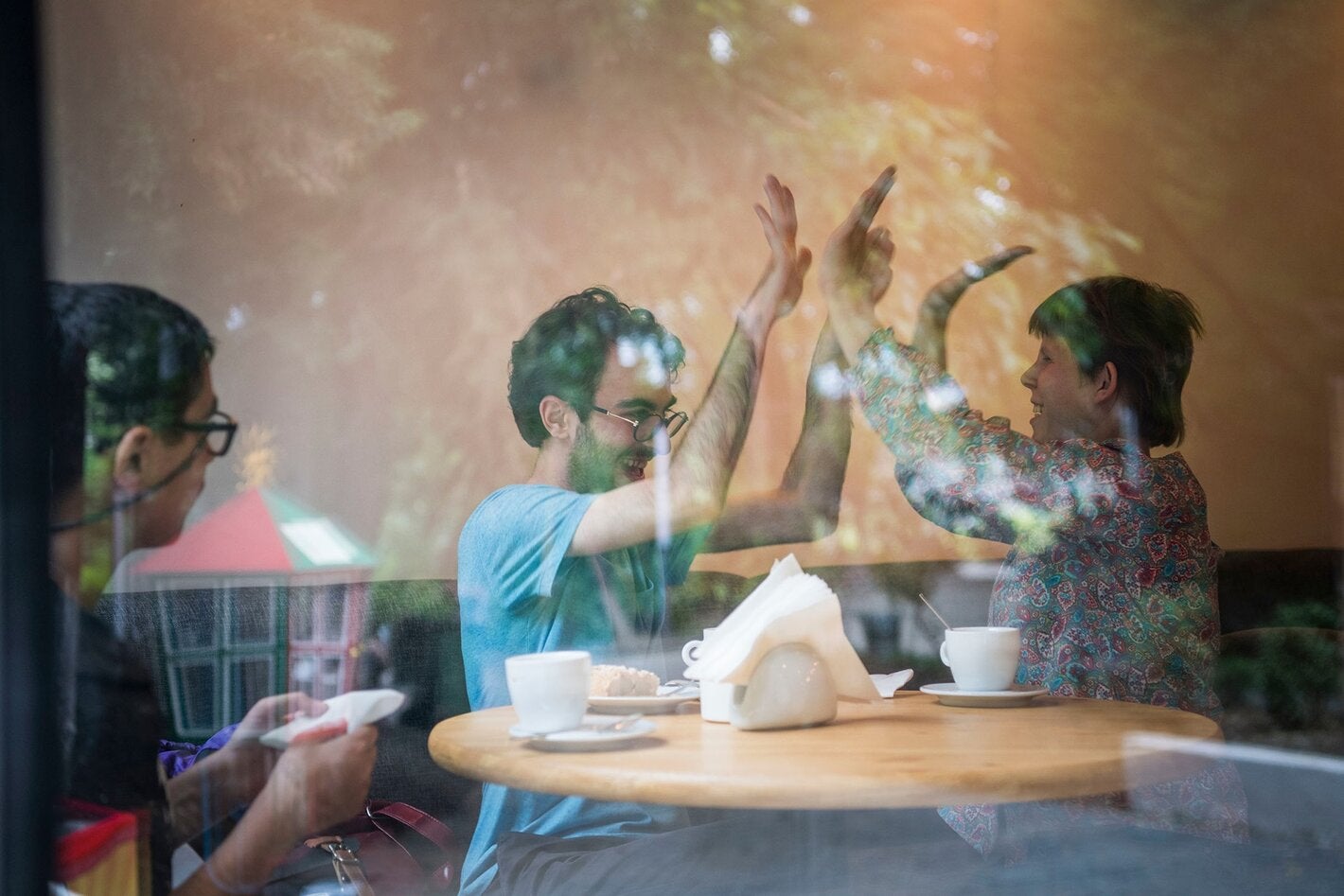 A man and a woman sitting next to a coffee table shake both hands with their arms up, while a third man watches the scene from the left