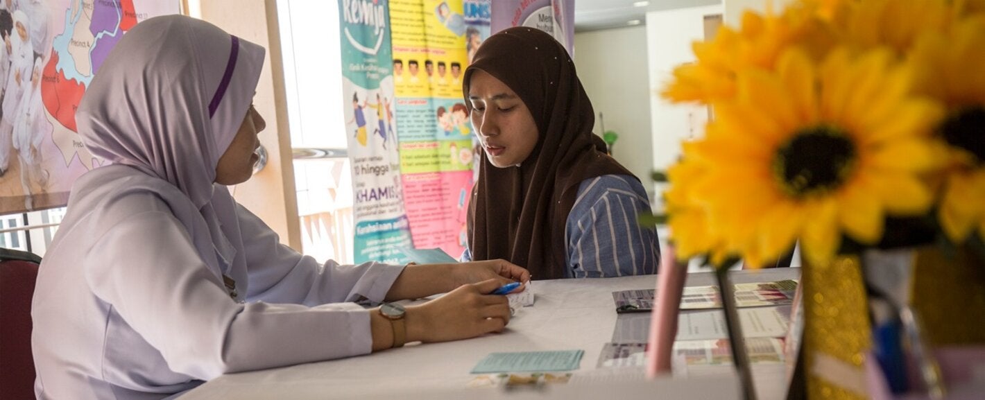 Photo of two women in health care facility; one is the patient, the other the health provider. Both have their heads covered with a scarf 